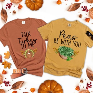 Puede incluir: Dos camisetas con juegos de palabras sobre el tema de Acci&oacute;n de Gracias. La camiseta naranja dice "Talk Turkey To Me" con un pavo de dibujos animados. La camiseta amarilla dice "Peas Be With You" con un taz&oacute;n de guisantes.