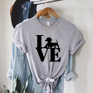 以下が含まれることがあります： 「LOVE」という言葉の上に立っているダックスフントの黒いシルエットが描かれた灰色のTシャツです。