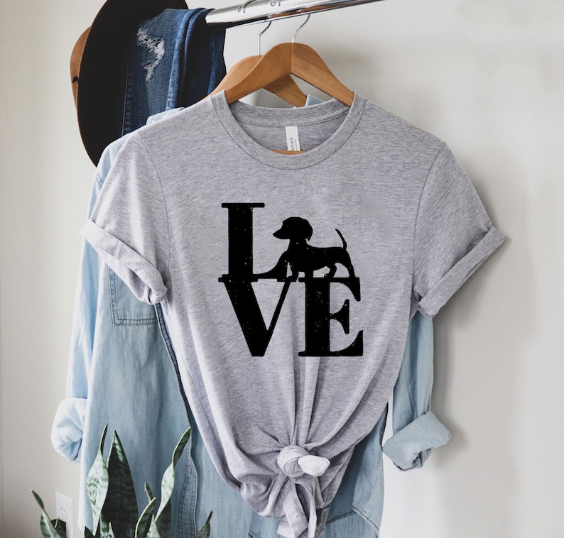 以下が含まれることがあります： 「LOVE」という言葉の上に立っているダックスフントの黒いシルエットが描かれた灰色のTシャツです。
