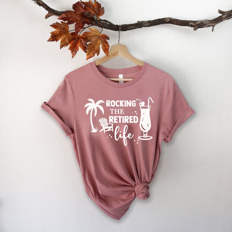 Op de afbeelding: Een roze T-shirt met een wit grafisch ontwerp met de tekst "Rocking the Retired Life" met een palmboom, een strandstoel en een cocktailglas.