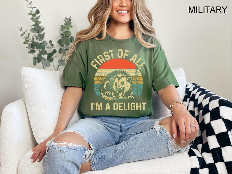 May include: A woman wearing a green t-shirt with a vintage-style graphic of an opossum and the text "First of all, I'm a delight".