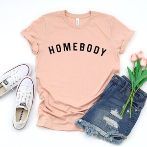 Puede incluir: Una camiseta de color melocot&oacute;n con la palabra "HOMEBODY" impresa en negro en la parte delantera. La camiseta se combina con pantalones cortos de mezclilla y zapatillas blancas.