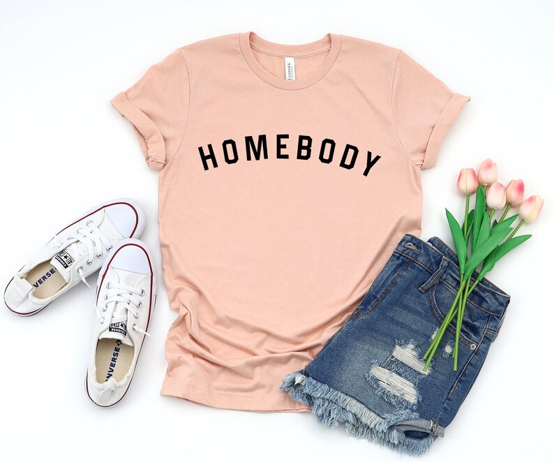 Puede incluir: Una camiseta de color melocot&oacute;n con la palabra "HOMEBODY" impresa en negro en la parte delantera. La camiseta se combina con pantalones cortos de mezclilla y zapatillas blancas.