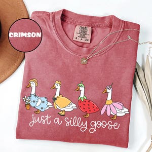 May include: Crimson Comfort Colors t-shirt featuring a whimsical illustration of four geese dressed in various outfits. The phrase "just a silly goose" is written in cursive below the geese. A gold necklace is draped across the shirt.