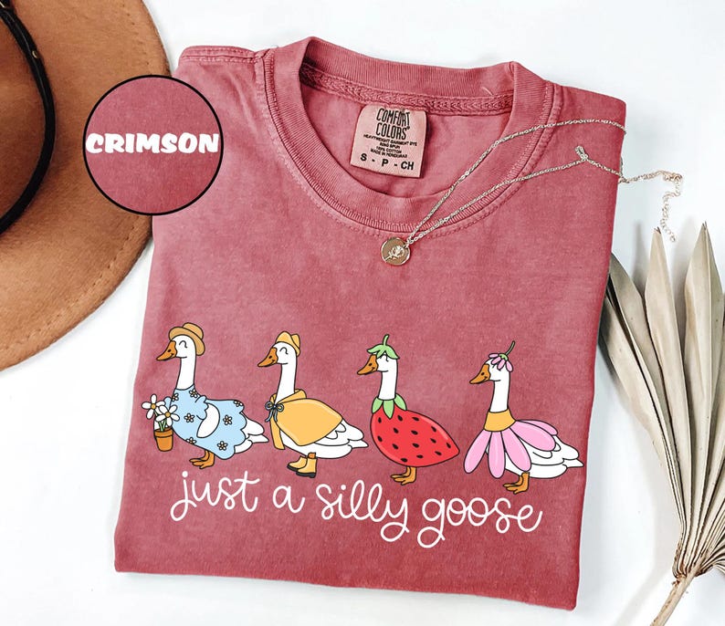 May include: Crimson Comfort Colors t-shirt featuring a whimsical illustration of four geese dressed in various outfits. The phrase "just a silly goose" is written in cursive below the geese. A gold necklace is draped across the shirt.