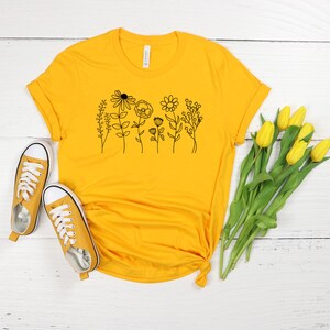 Pu&ograve; includere: Una maglietta gialla con un disegno di fiori in nero sul davanti. La maglietta &egrave; appoggiata su una superficie di legno bianco con un paio di scarpe da ginnastica gialle e un mazzo di tulipani gialli.