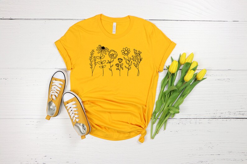 Pu&ograve; includere: Una maglietta gialla con un disegno di fiori in nero sul davanti. La maglietta &egrave; appoggiata su una superficie di legno bianco con un paio di scarpe da ginnastica gialle e un mazzo di tulipani gialli.