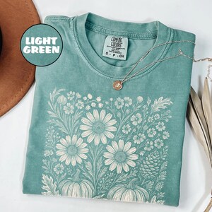May include: Light green t-shirt with a floral design featuring large daisies and pumpkins. The shirt has a crew neck and short sleeves. A silver necklace with a pendant is draped across the shirt. The shirt is labeled "Comfort Colors".