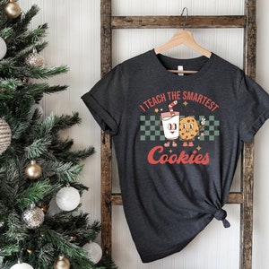 May include: A dark gray t-shirt with a retro design featuring a cartoon glass of milk and a cookie. The text on the shirt reads "I teach the smartest cookies".