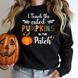 May include: Black sweatshirt with the text "I Teach the cutest PUMPKINS in the Patch." The word "PUMPKINS" is in orange, green, and patterned letters. An orange pumpkin graphic is below the text. A glittery orange pumpkin is on the left.