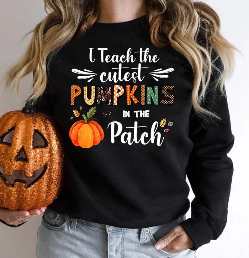 May include: Black sweatshirt with the text "I Teach the cutest PUMPKINS in the Patch." The word "PUMPKINS" is in orange, green, and patterned letters. An orange pumpkin graphic is below the text. A glittery orange pumpkin is on the left.