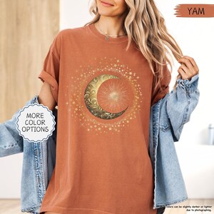 May include: A terracotta-colored t-shirt featuring a gold crescent moon and sun design surrounded by stars. The shirt has rolled-up sleeves and the text "MORE COLOR OPTIONS" is visible. The word "YAM" is also visible.