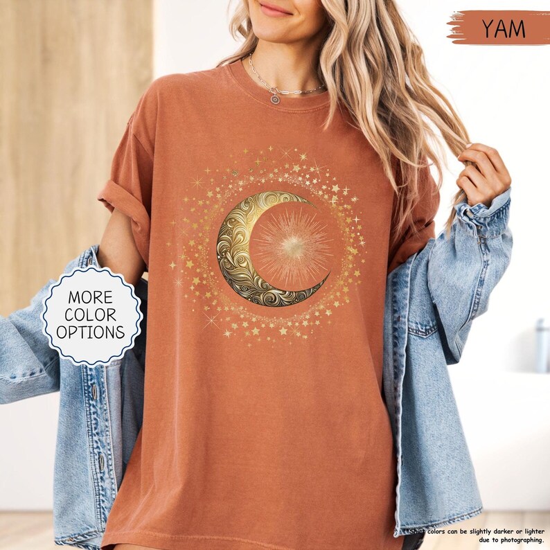 May include: A terracotta-colored t-shirt featuring a gold crescent moon and sun design surrounded by stars. The shirt has rolled-up sleeves and the text "MORE COLOR OPTIONS" is visible. The word "YAM" is also visible.
