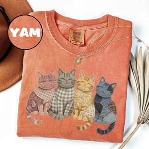 May include: Orange Comfort Colors t-shirt featuring four smiling cat designs with patchwork patterns. The shirt has a crew neck and short sleeves. The word "YAM" is in a circle on the upper left.