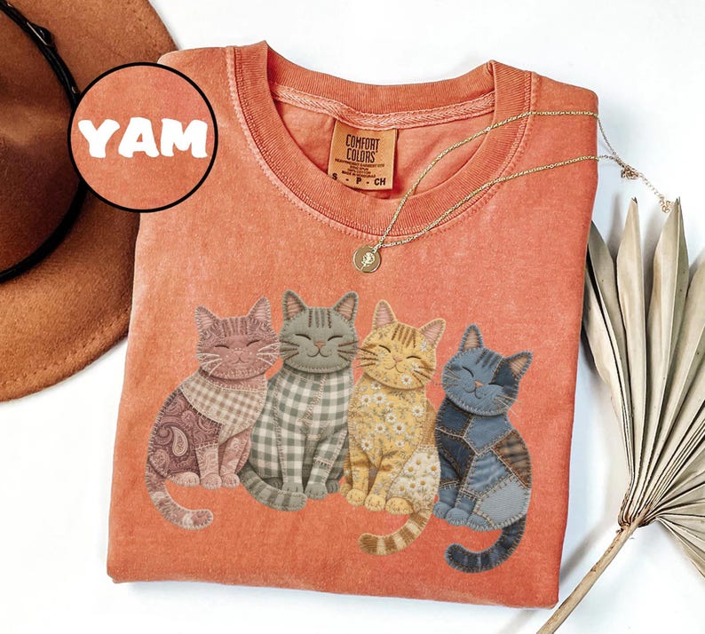 May include: Orange Comfort Colors t-shirt featuring four smiling cat designs with patchwork patterns. The shirt has a crew neck and short sleeves. The word "YAM" is in a circle on the upper left.