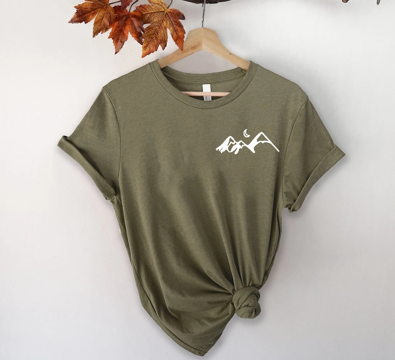 K&ouml;nnte beinhalten: Ein gr&uuml;nes T-Shirt mit kurzen &Auml;rmeln und einem wei&szlig;en Berg- und Halbmondmotiv auf der Vorderseite.