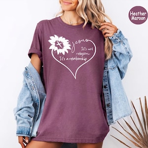 May include: A heather maroon t-shirt featuring a white graphic of a sunflower with a cross in the center, enclosed within a heart outline. The text reads: Jesus, It's not religion... It's a relationship. The shirt is worn with a light blue denim jacket.