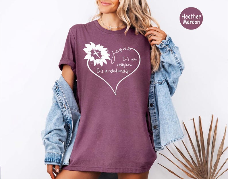 May include: A heather maroon t-shirt featuring a white graphic of a sunflower with a cross in the center, enclosed within a heart outline. The text reads: Jesus, It's not religion... It's a relationship. The shirt is worn with a light blue denim jacket.