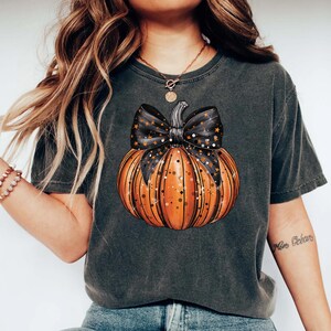 May include: A black t-shirt featuring a graphic of an orange pumpkin with a black bow and gold glitter. The bow has black stars on it.