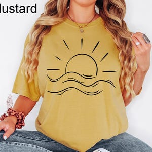 Puede incluir: Camiseta de color mostaza con un gr&aacute;fico negro de un sol sobre olas. La palabra "Mustard" est&aacute; impresa en negro en la esquina superior izquierda. La camiseta es una parte superior informal de manga corta.