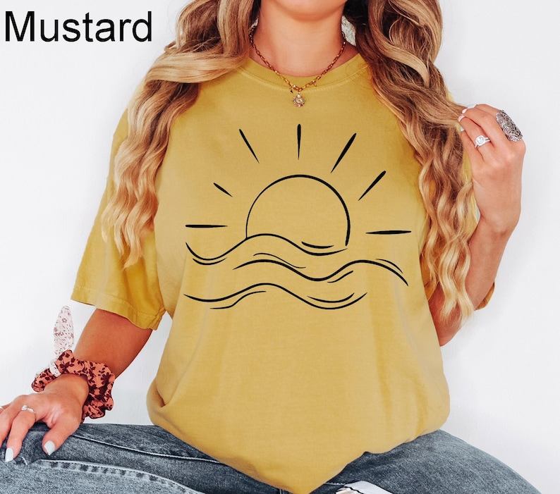 Puede incluir: Camiseta de color mostaza con un gr&aacute;fico negro de un sol sobre olas. La palabra "Mustard" est&aacute; impresa en negro en la esquina superior izquierda. La camiseta es una parte superior informal de manga corta.