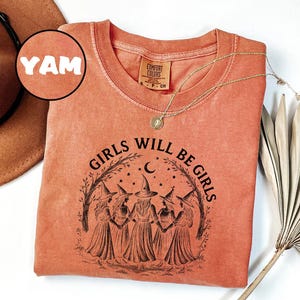 K&ouml;nnte beinhalten: Korallenfarbenes Langarmshirt mit dem Text "GIRLS WILL BE GIRLS" und einer Grafik von f&uuml;nf Hexen. Das Shirt hat einen Rundhalsausschnitt und ein kleines Etikett. Eine goldene Halskette mit Anh&auml;nger ist auf dem Shirt drapiert.