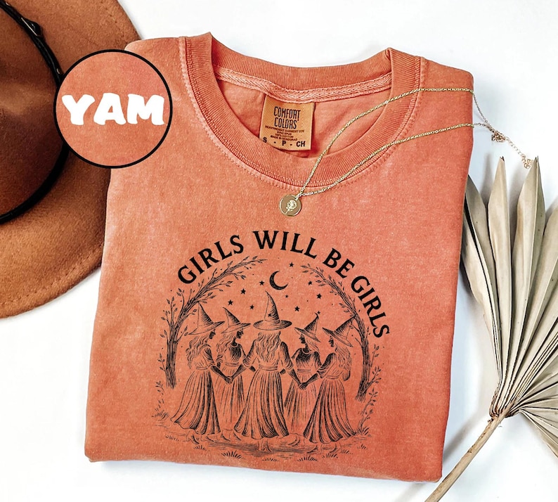 K&ouml;nnte beinhalten: Korallenfarbenes Langarmshirt mit dem Text "GIRLS WILL BE GIRLS" und einer Grafik von f&uuml;nf Hexen. Das Shirt hat einen Rundhalsausschnitt und ein kleines Etikett. Eine goldene Halskette mit Anh&auml;nger ist auf dem Shirt drapiert.