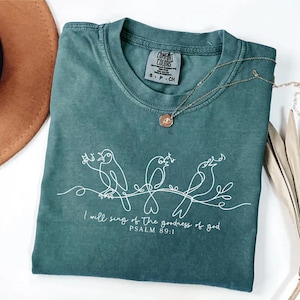 May include: A teal Comfort Colors t-shirt with a white line drawing of three birds on a branch. The text "I will sing of the goodness of god PSALM 89:1" is below the birds. A gold necklace with a small pendant is on the shirt.