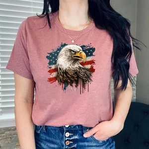 May include: A pink t-shirt with a graphic of a bald eagle with an American flag in the background. The eagle is facing right and has a white head and neck with brown feathers. The flag is red, white, and blue with white stars on a blue field.