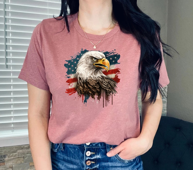 May include: A pink t-shirt with a graphic of a bald eagle with an American flag in the background. The eagle is facing right and has a white head and neck with brown feathers. The flag is red, white, and blue with white stars on a blue field.