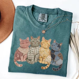 May include: Teal Comfort Colors t-shirt featuring four smiling cat illustrations with patchwork patterns in shades of pink, gray, yellow, and blue. A gold necklace with a pendant is draped across the shirt.