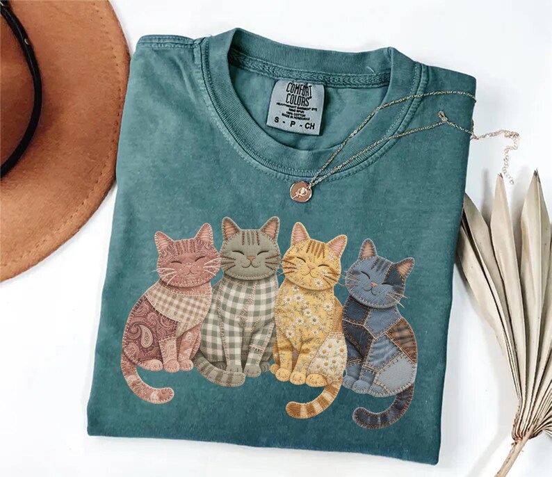 May include: Teal Comfort Colors t-shirt featuring four smiling cat illustrations with patchwork patterns in shades of pink, gray, yellow, and blue. A gold necklace with a pendant is draped across the shirt.