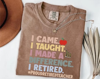 May include: A brown Comfort Colors t-shirt with the text "I CAME. I TAUGHT. I MADE A DIFFERENCE. I RETIRED." and a red apple graphic. A silver necklace with a pendant is draped across the shirt. The hashtag #PROUDRETIREDTEACHER is also visible.
