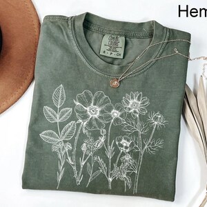 May include: Hemp-colored t-shirt with a white floral design. The shirt has a crew neck and short sleeves. A silver necklace with a pendant rests on the shirt. The tag reads "Comfort Colors".