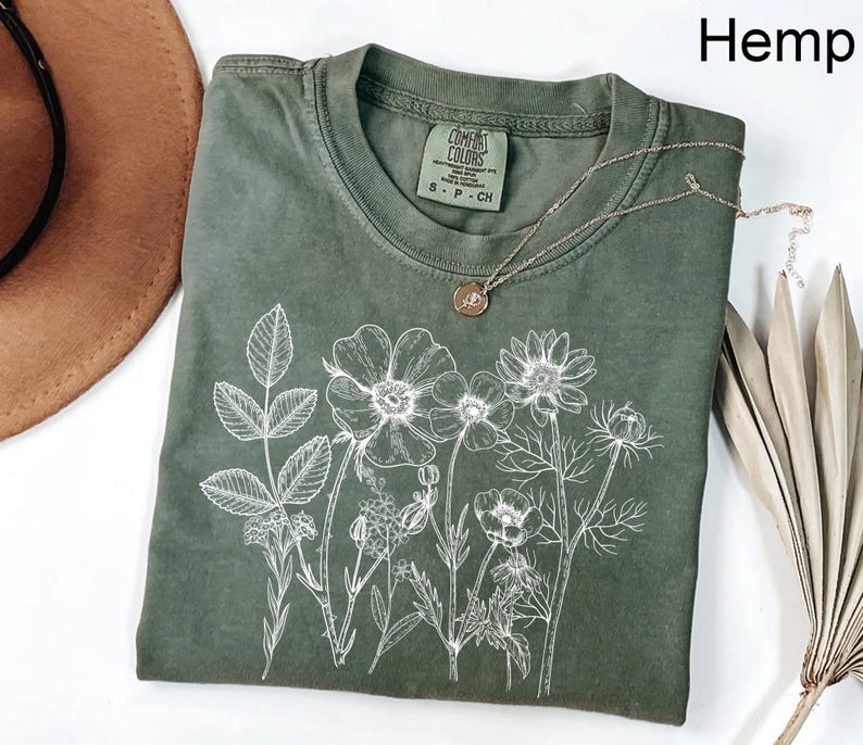 May include: Hemp-colored t-shirt with a white floral design. The shirt has a crew neck and short sleeves. A silver necklace with a pendant rests on the shirt. The tag reads "Comfort Colors".