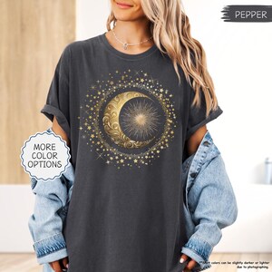 May include: Dark gray t-shirt featuring a gold crescent moon and sun design surrounded by gold stars. The shirt has rolled-up sleeves and the text "MORE COLOR OPTIONS" is visible. The word "PEPPER" is also visible.
