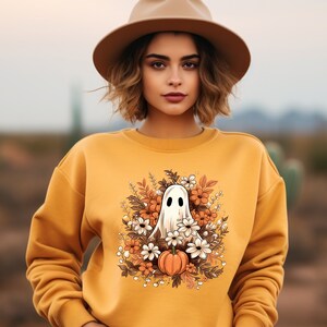 May include: Mustard yellow sweatshirt featuring a Halloween-themed graphic. The graphic includes a white ghost, orange pumpkin, and various flowers in shades of orange, brown, and white. The sweatshirt is a solid color and has a crew neck.