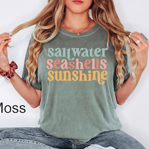 May include: Moss green t-shirt with the words "saltwater seashells sunshine" in colorful, retro-style lettering. The shirt is a relaxed fit and appears to be made of soft material. The word "Moss" is printed in black at the bottom left.