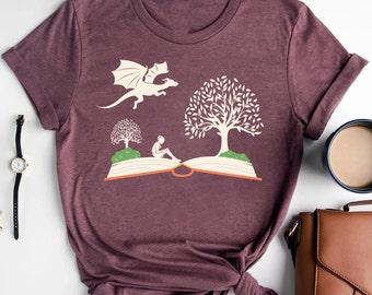 May include: A maroon t-shirt with a graphic of a dragon flying over an open book with a person sitting on the book and two trees.