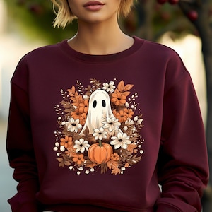 May include: Burgundy sweatshirt featuring a Halloween-themed design. The graphic includes a white ghost, orange pumpkin, and various flowers in shades of orange, brown, and white. The sweatshirt is a solid burgundy color.