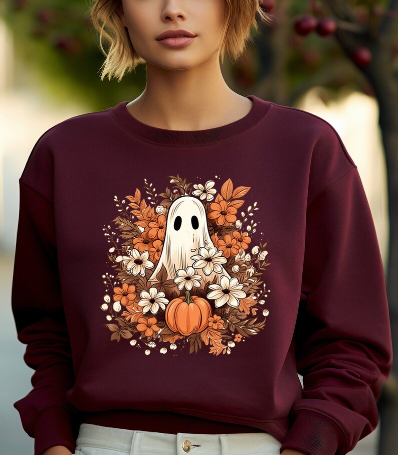 May include: Burgundy sweatshirt featuring a Halloween-themed design. The graphic includes a white ghost, orange pumpkin, and various flowers in shades of orange, brown, and white. The sweatshirt is a solid burgundy color.