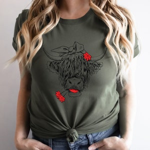 May include: A green t-shirt with a black and white illustration of a Highland cow wearing a red flower headband and holding a red flower in its mouth.