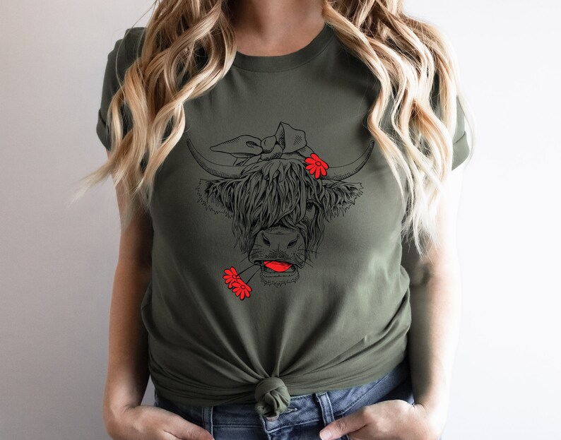 May include: A green t-shirt with a black and white illustration of a Highland cow wearing a red flower headband and holding a red flower in its mouth.