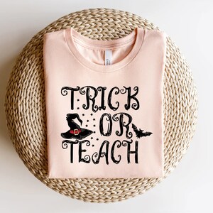 Puede incluir: Una camiseta rosa claro con las palabras "TRICK OR TEACH" en escritura negra. El dise&ntilde;o incluye un sombrero de bruja negro con estrellas y un murci&eacute;lago. La camiseta est&aacute; doblada sobre una superficie tejida de color marr&oacute;n claro.