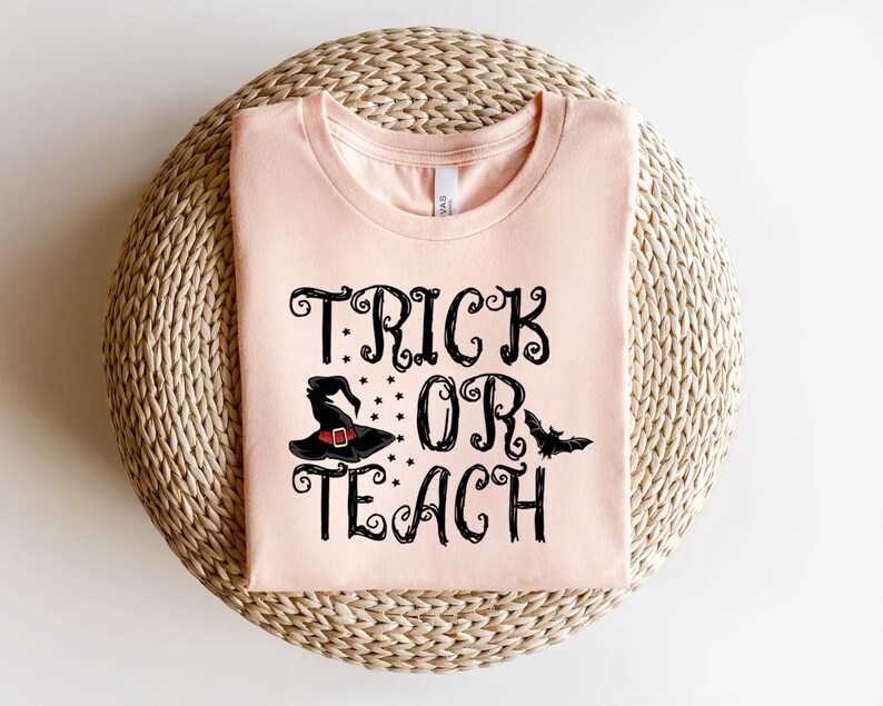 Puede incluir: Una camiseta rosa claro con las palabras "TRICK OR TEACH" en escritura negra. El dise&ntilde;o incluye un sombrero de bruja negro con estrellas y un murci&eacute;lago. La camiseta est&aacute; doblada sobre una superficie tejida de color marr&oacute;n claro.