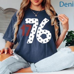Puede incluir: Una camiseta azul marino con un gr&aacute;fico blanco y rojo del n&uacute;mero 1776 hecho de estrellas. La camiseta la lleva puesta una persona sentada en una cama blanca con pantalones vaqueros.