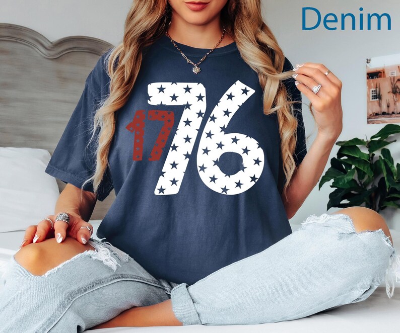 Puede incluir: Una camiseta azul marino con un gr&aacute;fico blanco y rojo del n&uacute;mero 1776 hecho de estrellas. La camiseta la lleva puesta una persona sentada en una cama blanca con pantalones vaqueros.