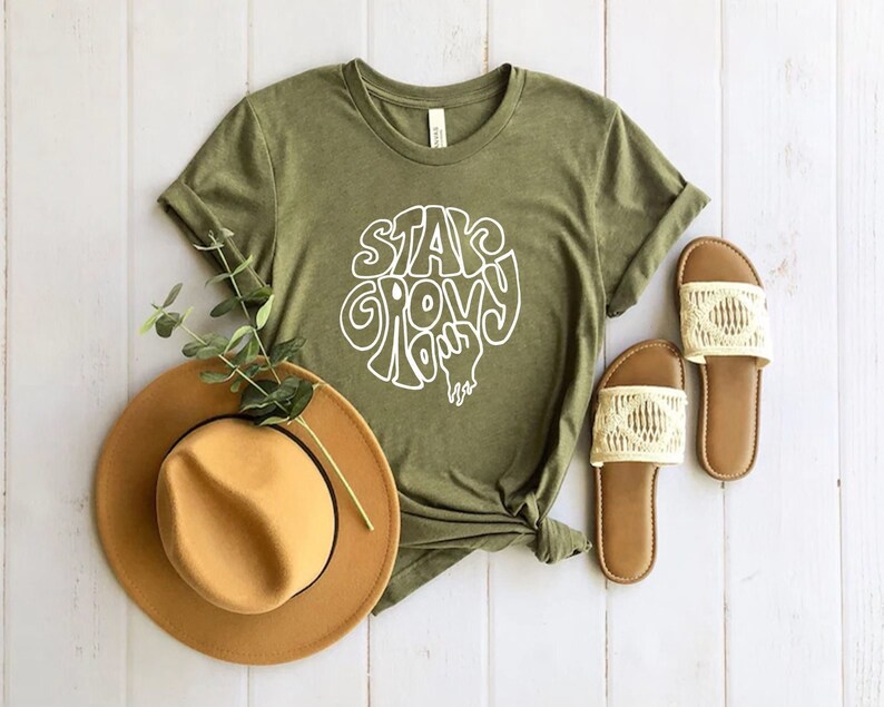 May include: A green t-shirt with the text "Stay Groovy" in white lettering. The shirt is laid out on a white wooden surface with a brown hat and white sandals.