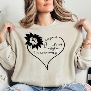 May include: A sand-colored crewneck sweatshirt featuring a black graphic of a sunflower with a cross in the center, forming part of a heart shape. The text reads: Jesus It's not religion... It's a relationship. The sweatshirt has long sleeves.