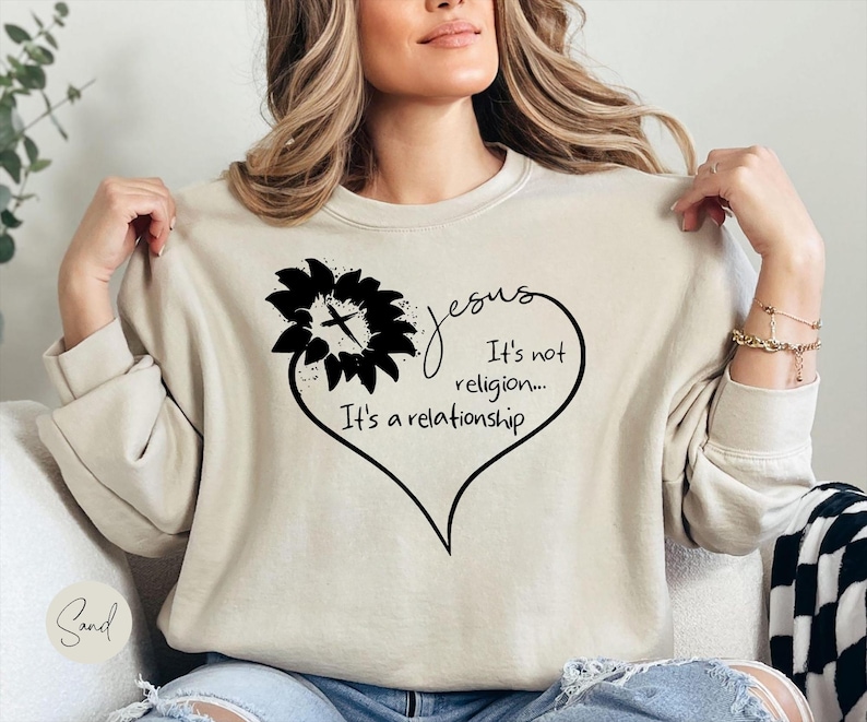 May include: A sand-colored crewneck sweatshirt featuring a black graphic of a sunflower with a cross in the center, forming part of a heart shape. The text reads: Jesus It's not religion... It's a relationship. The sweatshirt has long sleeves.